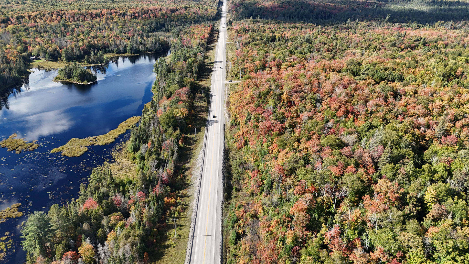 Aerial view of a straight two-lane highway running through a dense forest with vibrant autumn foliage. On the left side of the road, there is a large blue lake surrounded by trees, while the right side is covered with colorful forest in shades of red, orange, yellow, and green. A few vehicles are visible traveling along the highway under a clear sky.